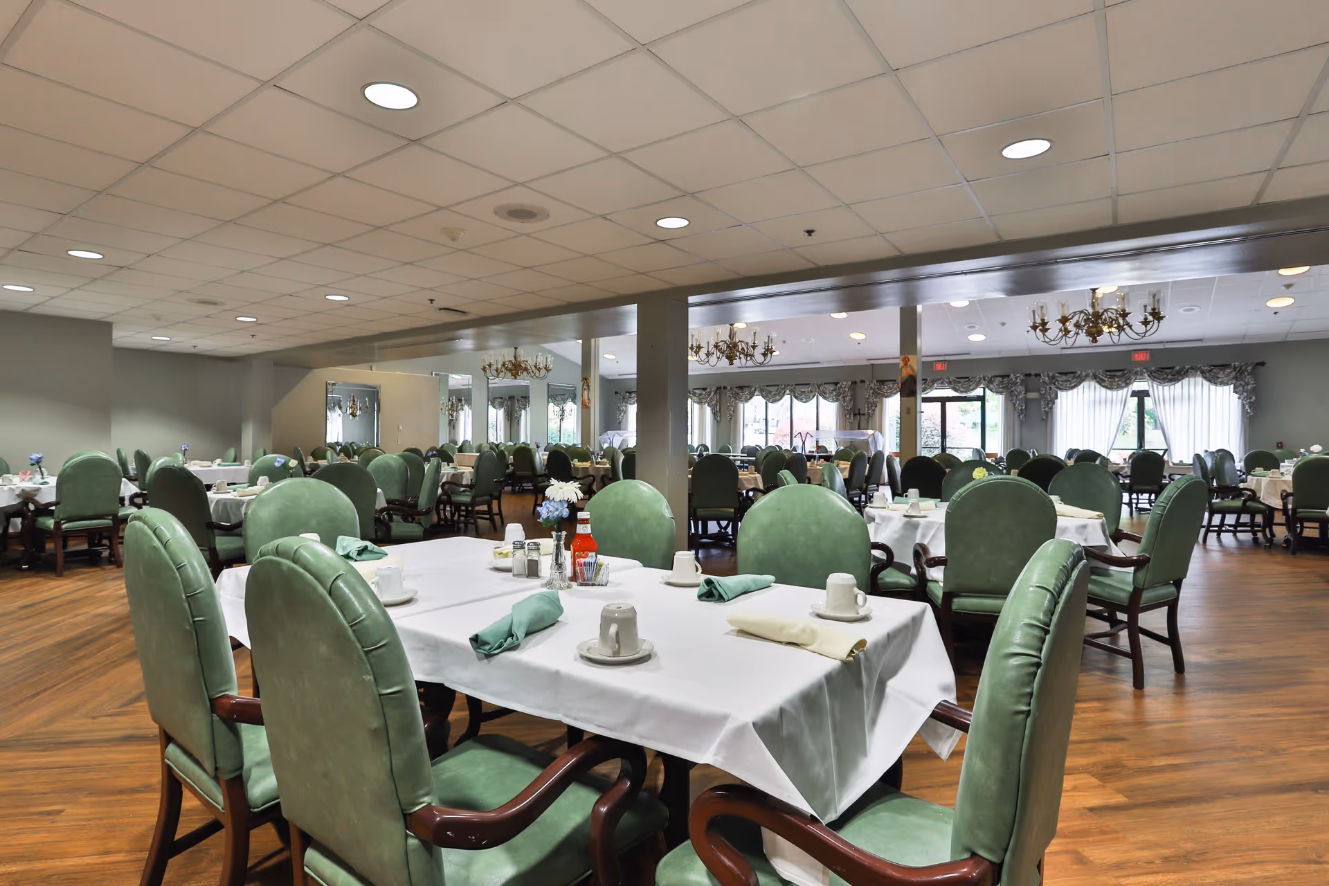 A spacious dining room with multiple tables covered in white tablecloths, each set with cups, saucers, napkins, and condiments. The room features green upholstered chairs, wooden flooring, chandeliers, and large windows with curtains allowing natural light to enter.