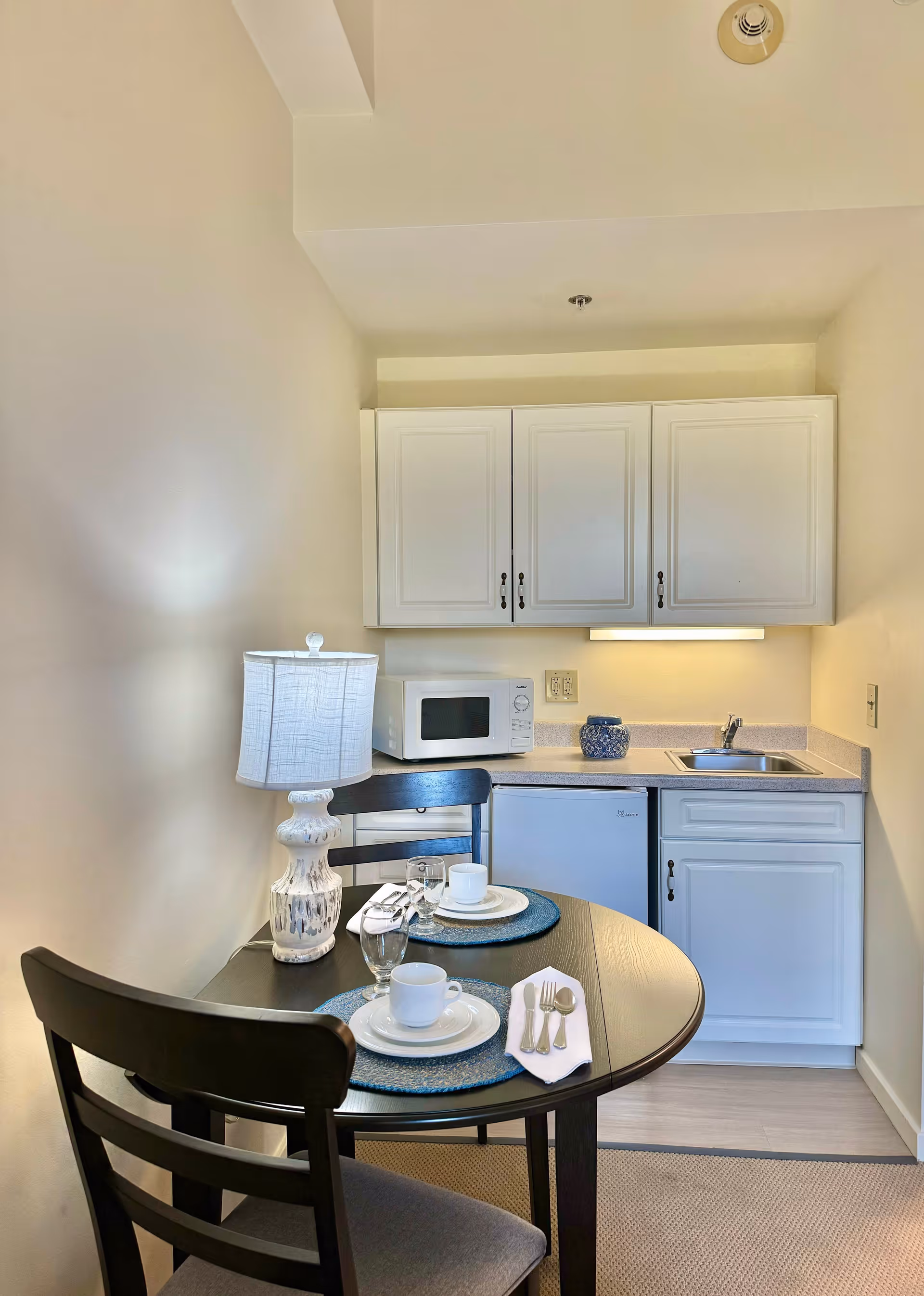 Small kitchenette with white cabinets, microwave, sink and mini-fridge beside a round dining table set for two with a lamp.