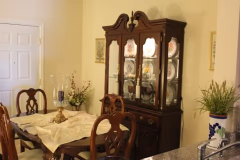 A dining room with a wooden dining table covered with a cream-colored tablecloth and surrounded by wooden chairs. Behind the table is a wooden china cabinet displaying decorative plates. There are floral arrangements on the table and beside the cabinet. The room has light-colored walls and a door is visible in the background.