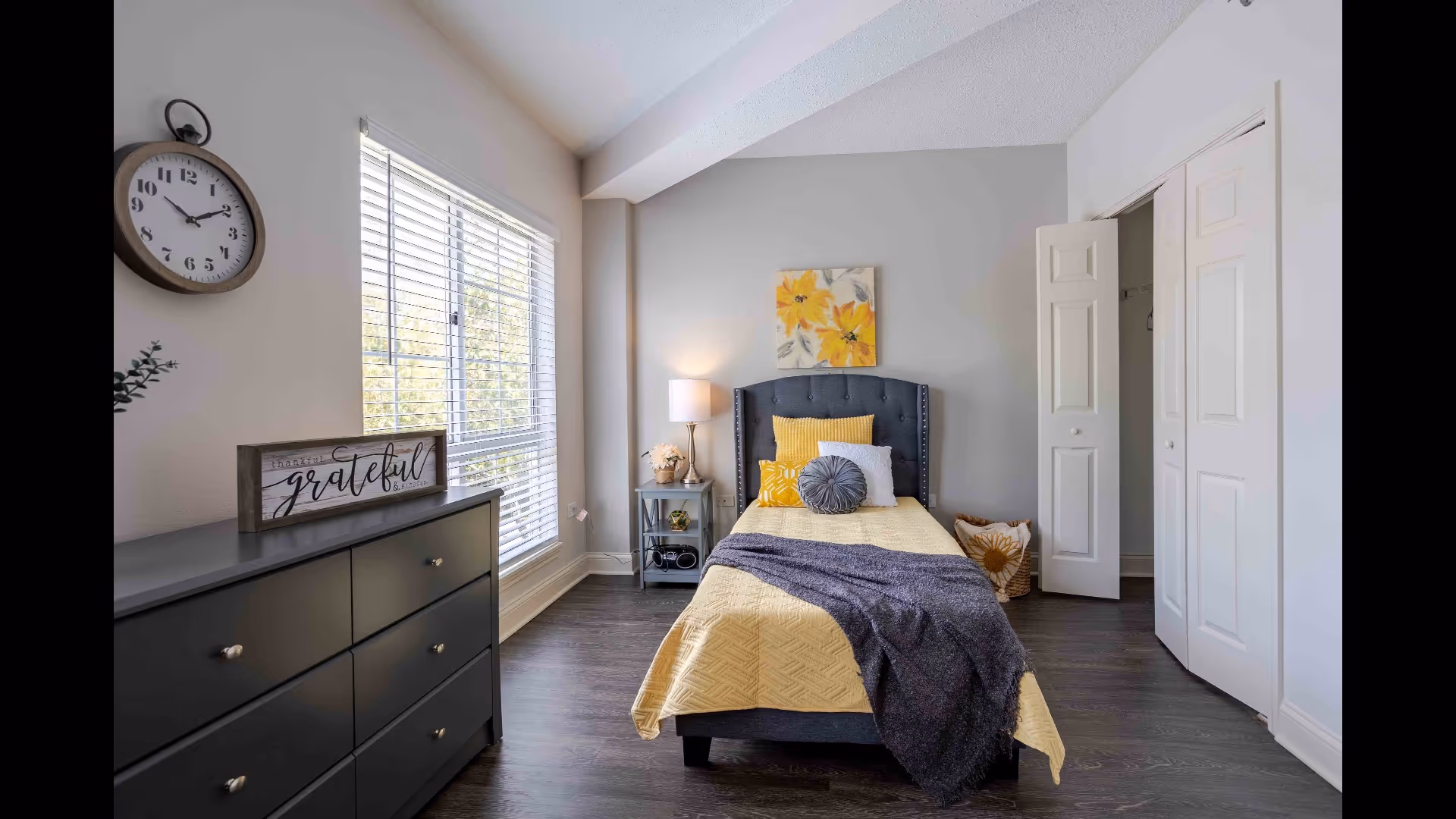 A bright bedroom with a single bed featuring a yellow bedspread and multiple pillows, including a round gray one. There is a dark gray dresser with a decorative sign that says 'grateful' and a round wall clock above it. A window with white blinds lets in natural light. Next to the bed is a small nightstand with a lamp and decorative items. The room has dark wood flooring and light-colored walls, with a closet door open on the right side.