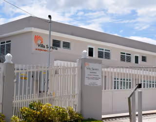Exterior front of the Villa Serena building behind a white security fence and gate under a partly cloudy sky.