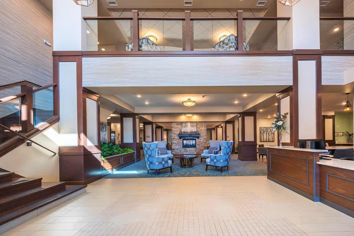 Spacious and well-lit senior living facility lobby with a seating area featuring four blue patterned armchairs around a round wooden table in front of a stone fireplace. To the right is a wooden reception desk with a computer and a vase with flowers. A staircase with glass and wood railing is on the left, and the upper level has a glass partition with additional seating visible.
