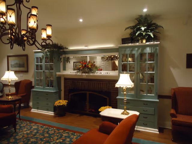 Cozy living room with a brick fireplace flanked by teal glass-front cabinets, upholstered chairs, lamps, and a floral centerpiece.