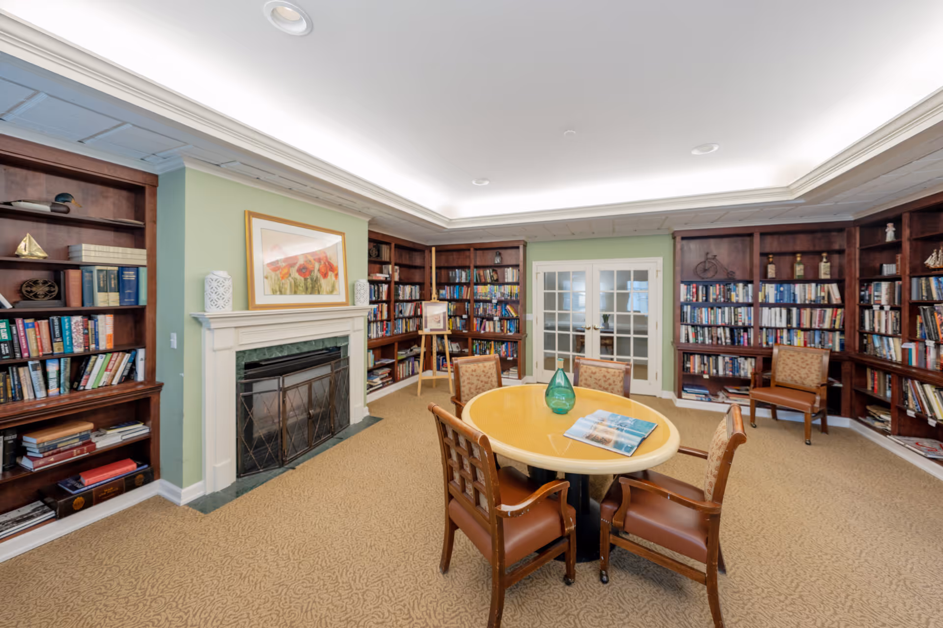 Cozy communal library room with built-in bookshelves, a fireplace, a round table with chairs, and French doors under recessed ceiling lighting.