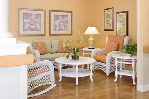 A cozy sitting area with white wicker furniture including a loveseat, two armchairs, and a rocking chair. The loveseat has light green cushions, and the chairs have orange cushions with patterned pillows. A white round coffee table and a matching side table hold decorative plants and a lamp. The walls are light yellow with framed floral artwork.