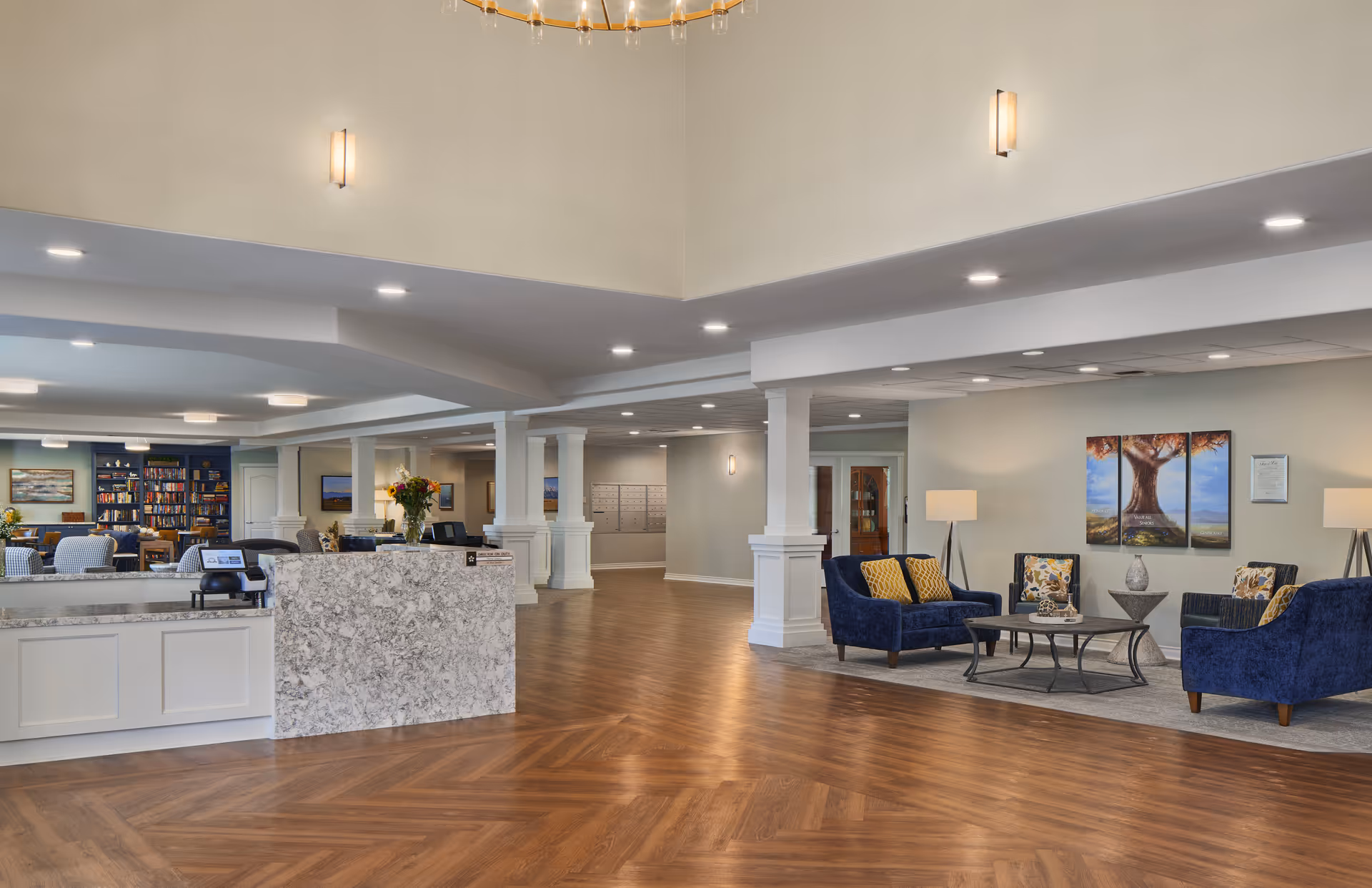 Spacious senior living facility lobby with a marble reception desk, wooden herringbone floor, blue upholstered seating area with yellow and floral cushions, modern lamps, and a bookshelf in the background.