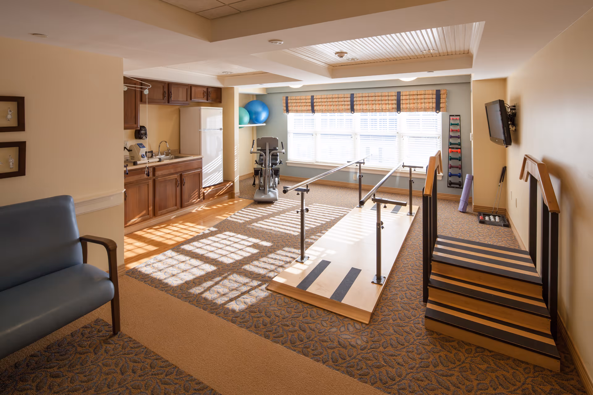 A well-lit physical therapy room with parallel bars for walking exercises, a small set of stairs with handrails, exercise balls, a stationary exercise machine, and a rack of colorful hand weights. The room has carpeted flooring, a blue armchair, wooden cabinets, a sink, and a refrigerator. A wall-mounted TV is visible on the right side.
