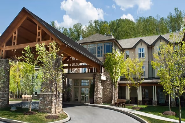 Exterior view of a senior living facility named Spring Mill with a covered entrance supported by stone pillars, surrounded by young trees and landscaping under a partly cloudy sky.