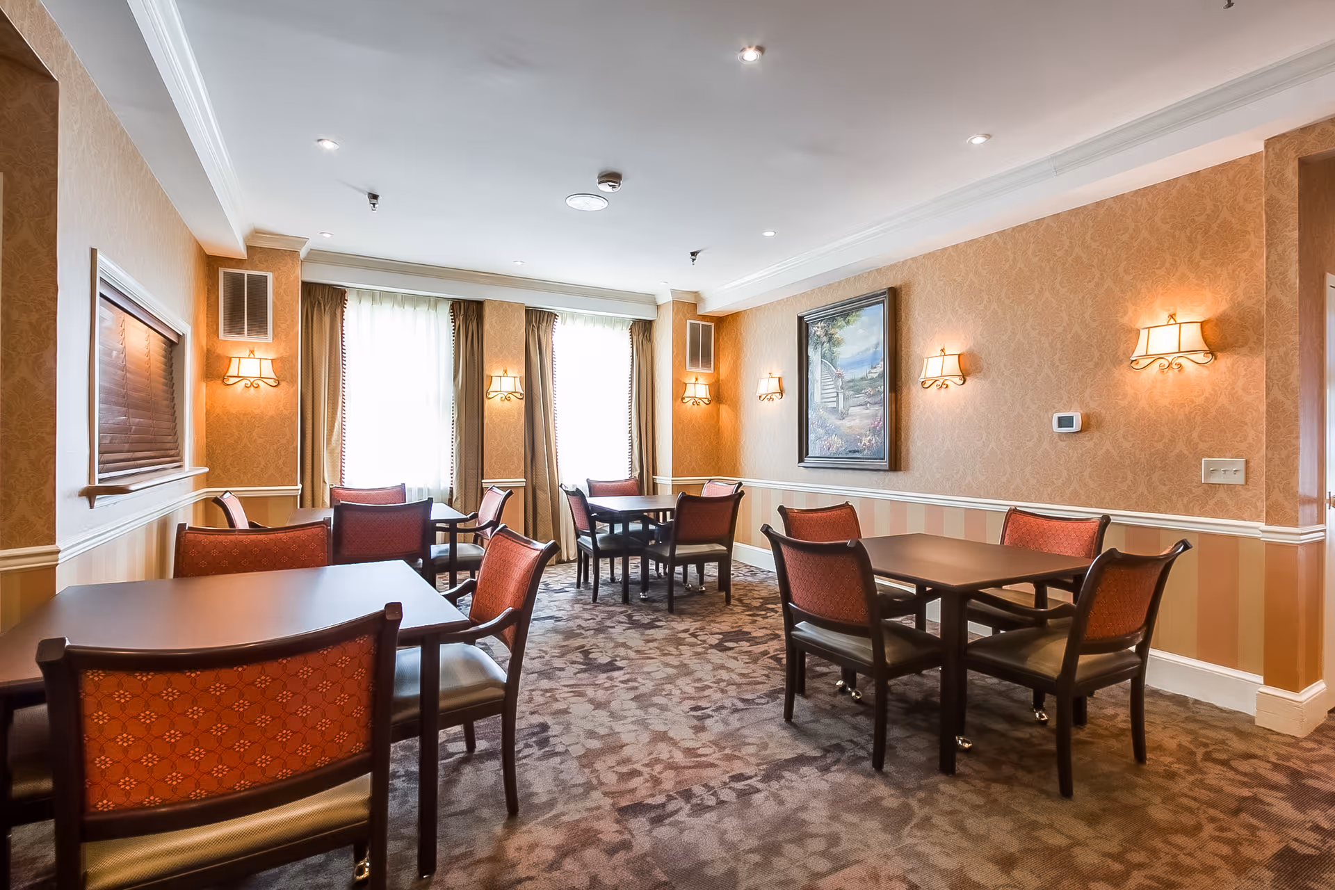 A cozy dining room in an assisted living facility featuring several square tables with red cushioned chairs. The room has patterned wallpaper, wall sconces with warm lighting, a framed painting, and large windows with curtains allowing natural light to enter. The carpet has a floral design, and the overall ambiance is warm and inviting.