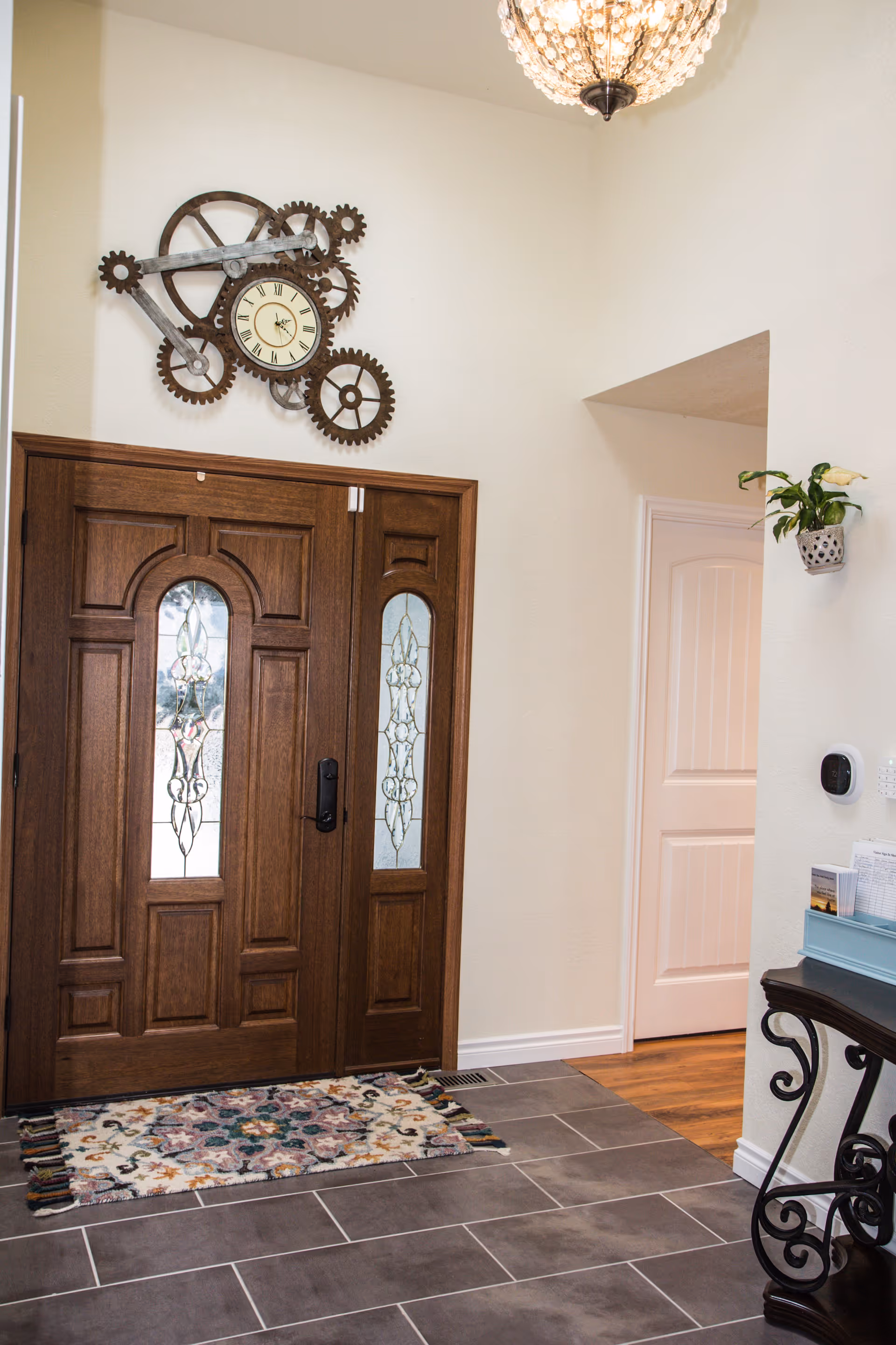 Interior view of an entryway with a wooden front door featuring decorative glass panels. Above the door is a wall clock designed with large metal gears. A colorful patterned rug is placed on the tiled floor in front of the door. To the right, there is a small table with a blue organizer and a potted plant hanging on the wall above it. A thermostat and a keypad are also visible on the wall.