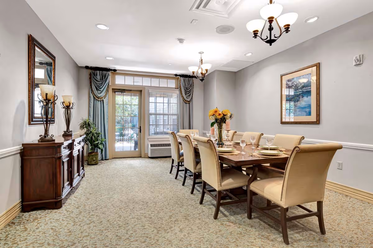 A formal dining room with a long wooden table set with place settings, upholstered chairs, chandeliers, a sideboard, and French doors with drapes.