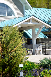 Covered front entrance of a light-colored building with a teal metal roof, white trim, outdoor chairs, and surrounding landscaping.