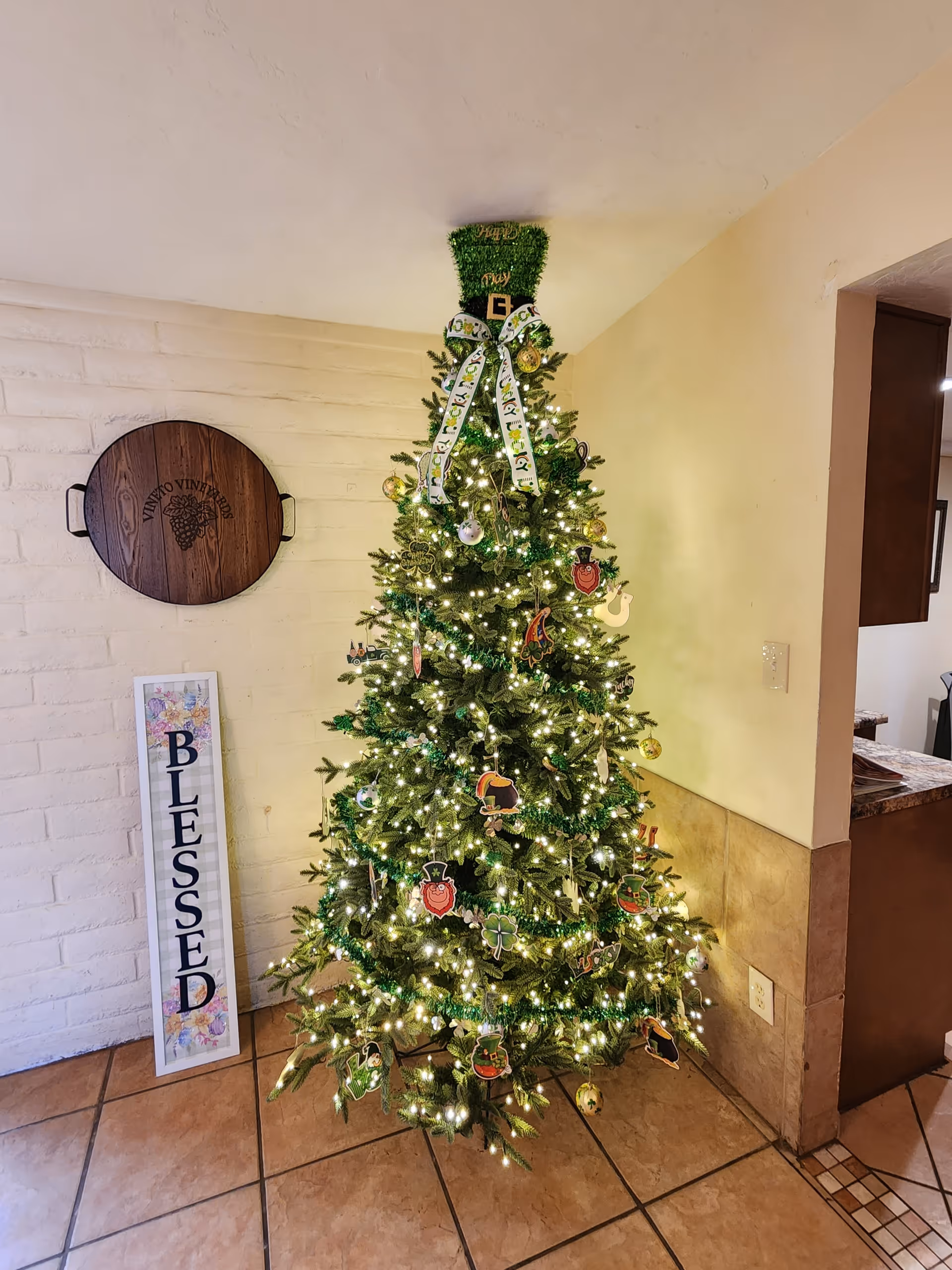 A decorated Christmas tree with white lights and various ornaments stands in the corner of a room with tiled floors and light-colored walls. To the left of the tree is a wooden round tray mounted on the wall and a vertical sign that reads 'BLESSED'.