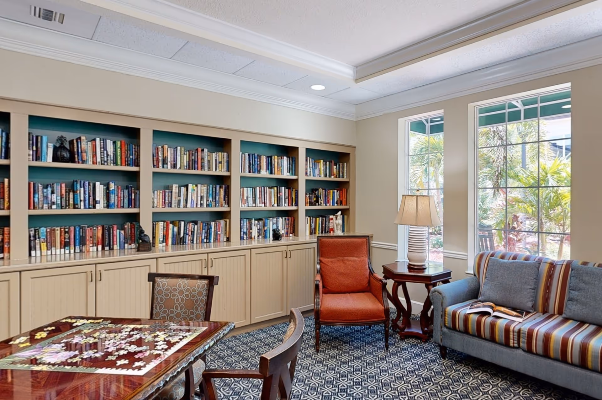 A cozy reading and activity room with built-in bookshelves filled with books, a table with a puzzle in progress, an upholstered armchair, a striped sofa with cushions, a side table with a lamp, and large windows showing greenery outside.