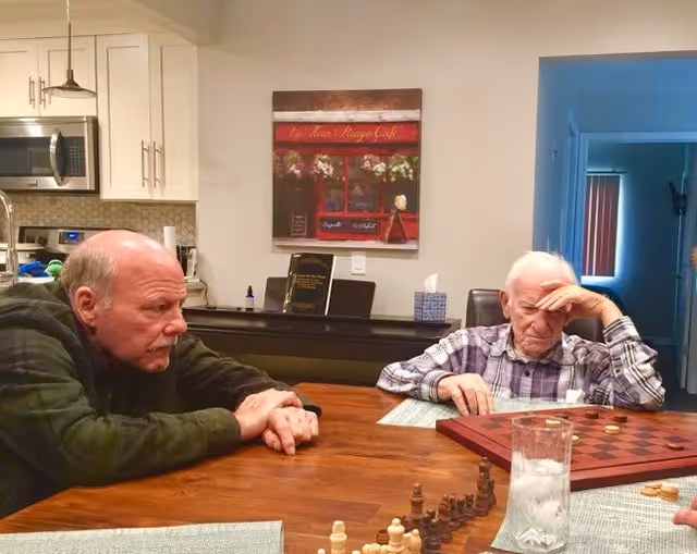 Two elderly men sitting at a wooden table in a kitchen or dining area, playing a board game. One man is leaning forward with his hands clasped, while the other is resting his head on his hand, concentrating on the game. The background shows kitchen cabinets, a microwave, and a framed picture on the wall.