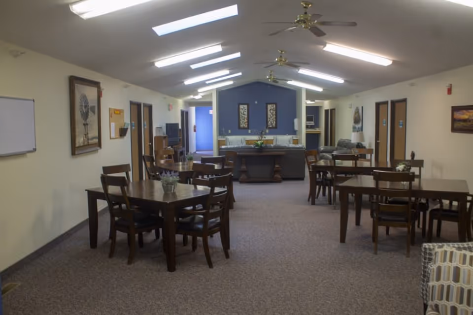 Interior view of a senior living facility common area with several wooden tables and chairs arranged on a carpeted floor. The room has a high ceiling with skylights and ceiling fans. At the far end, there is a seating area with sofas and framed artwork on a blue accent wall. Doors line the walls on both sides of the room.
