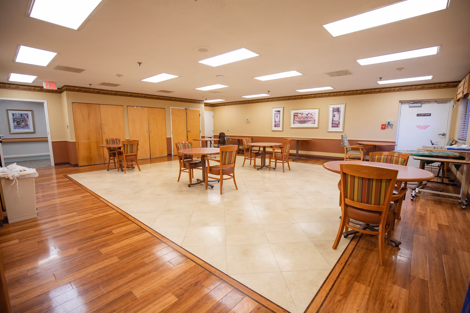 A spacious room with a mix of wooden and tiled flooring, featuring several round wooden tables each surrounded by four chairs with striped upholstery. The walls are beige with a brown trim and decorated with framed artwork. The ceiling has multiple rectangular fluorescent lights. There are several closed wooden doors along one wall and a white door labeled 'COURTYARD' on another wall. The room appears clean and well-lit.