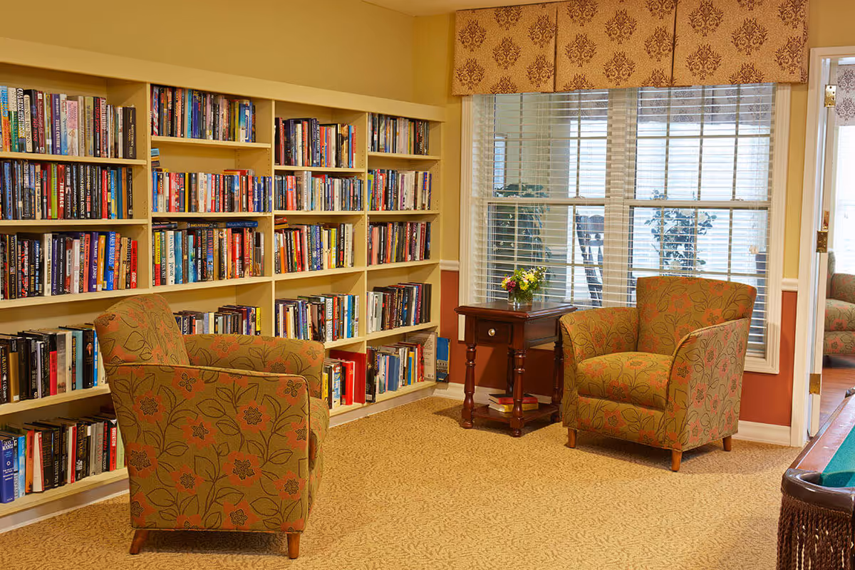 Cozy reading area with built-in bookshelves, two patterned armchairs, a small side table and a window with blinds.
