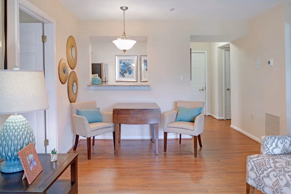 A cozy interior space featuring two beige armchairs with blue cushions positioned around a small wooden table. The room has light-colored walls and wood flooring. On the left side, there is a table with a blue lamp and a framed photo. Decorative round wall hangings are visible on the left wall. A pass-through window reveals part of a kitchen with a refrigerator and framed floral artwork.