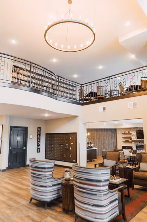 Interior view of a senior living facility lounge area with comfortable striped armchairs, wooden side tables, and a welcoming atmosphere. The space features a high ceiling with a large circular chandelier, a second-floor balcony with bookshelves, and a cozy seating area with armchairs and a fireplace in the background.