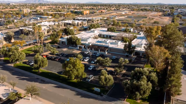 Aerial view of Chandler Post Acute and Rehabilitation facility showing a single-story building with a white roof surrounded by trees and parking lots filled with cars. The surrounding area includes residential neighborhoods and distant mountains under a clear sky.
