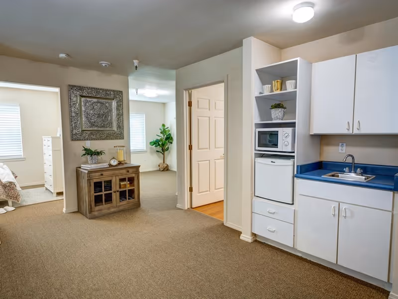 Interior view of a senior living facility unit showing a small kitchenette with white cabinets, a blue countertop, a sink, a microwave, and a mini refrigerator. Adjacent to the kitchenette is a hallway leading to a bedroom with a bed and dresser visible. The area is carpeted and decorated with a small cabinet, a plant, and a framed artwork on the wall.
