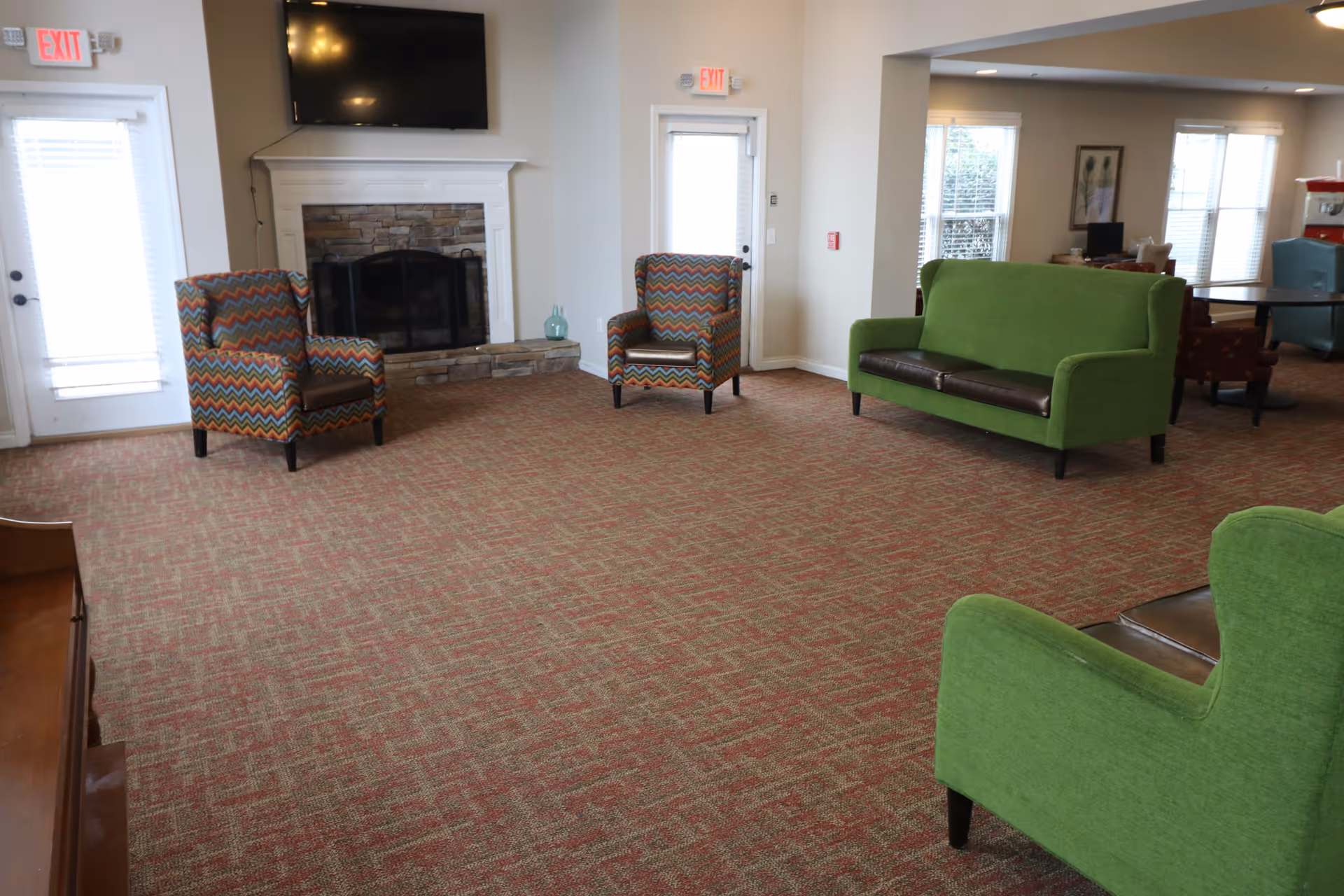Spacious senior living common room with green sofas, patterned armchairs, a fireplace and a wall-mounted TV.