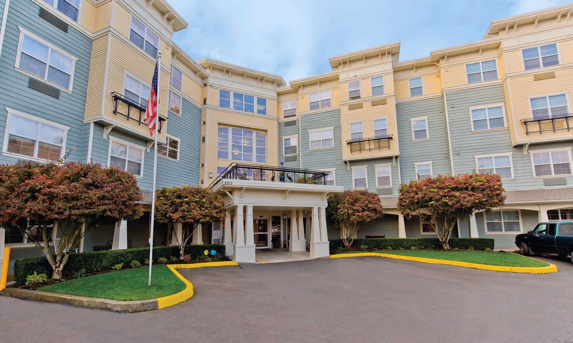 Front exterior view of a multi-story senior living facility building with green and beige siding, multiple windows, a covered entrance with white pillars, an American flag on a flagpole, and landscaped bushes and trees along the driveway.