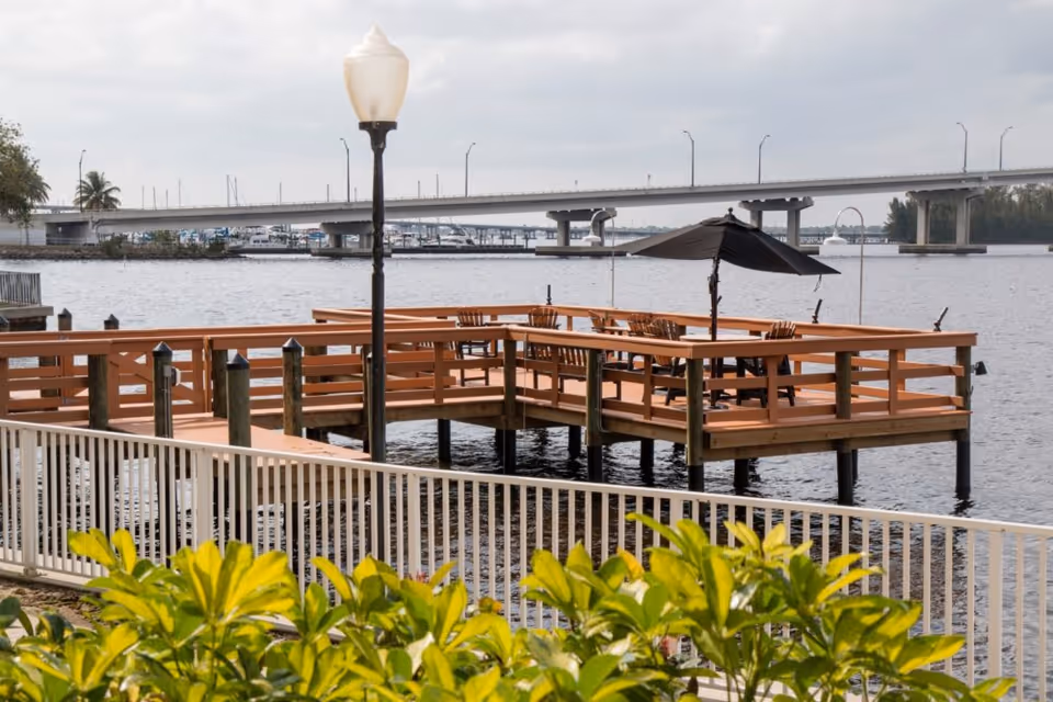 Wooden dock with seating area and umbrella extending over a body of water, with a bridge and boats visible in the background under a cloudy sky.