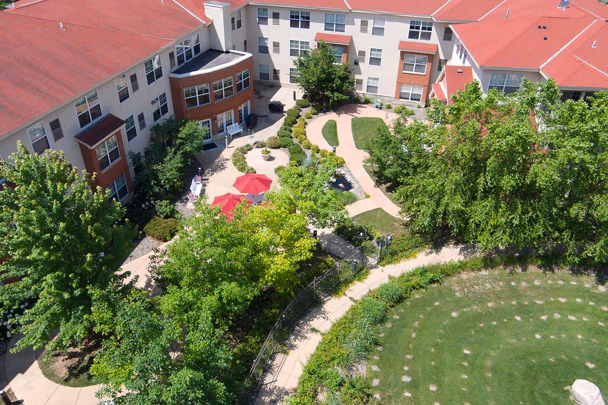 Aerial view of a landscaped courtyard with winding paths, trees, red patio umbrellas and the surrounding senior living building.