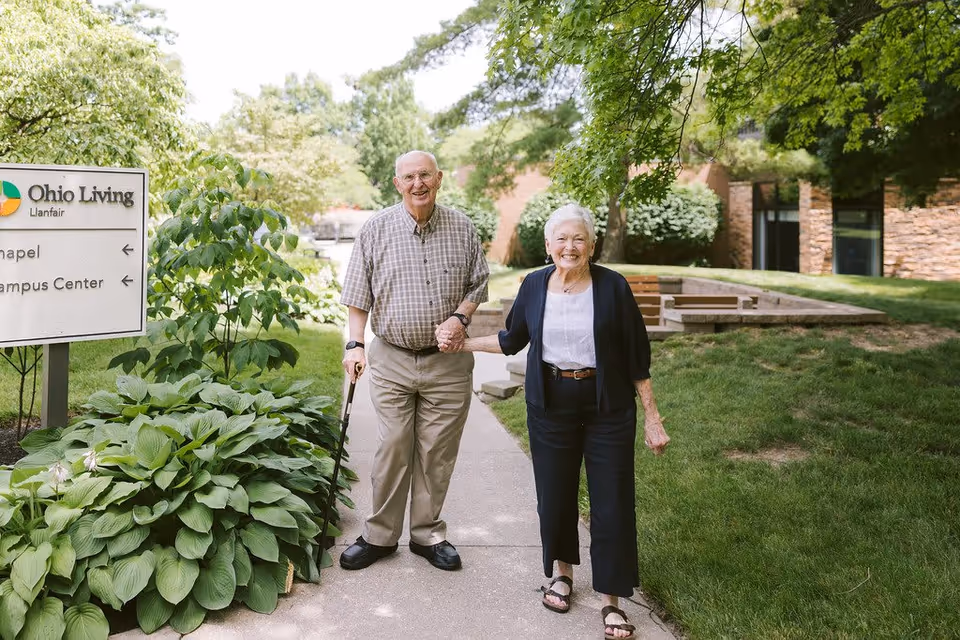 An elderly man and woman holding hands and smiling while standing on a sidewalk outside near greenery and a building. A sign nearby reads 'Ohio Living Llanfair' with arrows pointing to the Chapel and Campus Center.