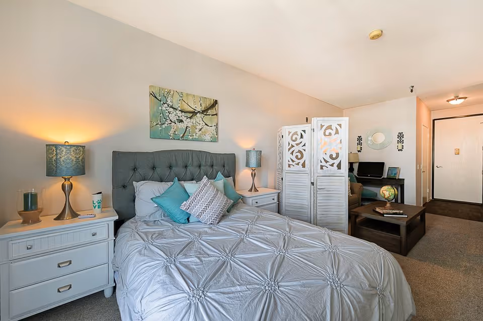 A cozy bedroom with a neatly made bed featuring a tufted gray headboard and decorative pillows in shades of teal and gray. On either side of the bed are white nightstands with matching lamps emitting a warm light. Above the bed hangs a painting of white blossoms on branches. A decorative white folding screen partially separates the sleeping area from a small sitting area with a coffee table, globe, and a computer desk against the far wall. The room has beige walls and carpeted flooring.