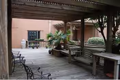 Outdoor covered patio area with wooden decking, benches, tables, and metal chairs. There are plants and shrubs along the edges and a brick wall in the background.