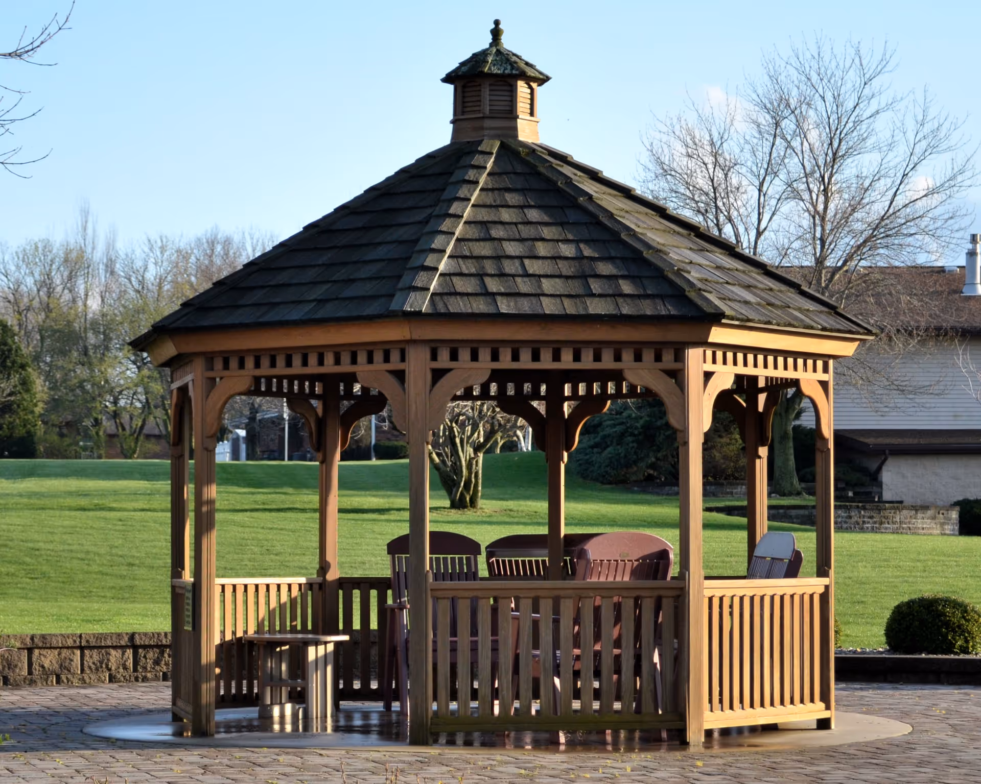 Wooden gazebo with seating on a paved area overlooking a grassy lawn and trees.
