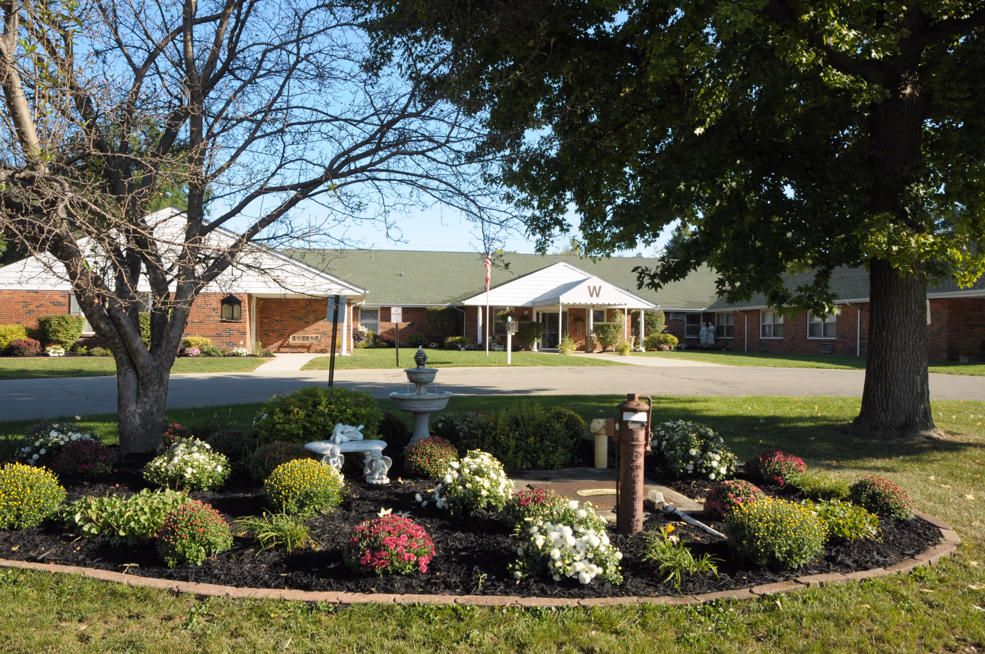 A landscaped garden area with colorful flowers, a small stone bench, and a birdbath in front of a single-story brick building with a green roof and a covered entrance marked with a large letter W. Trees provide shade around the garden and building.