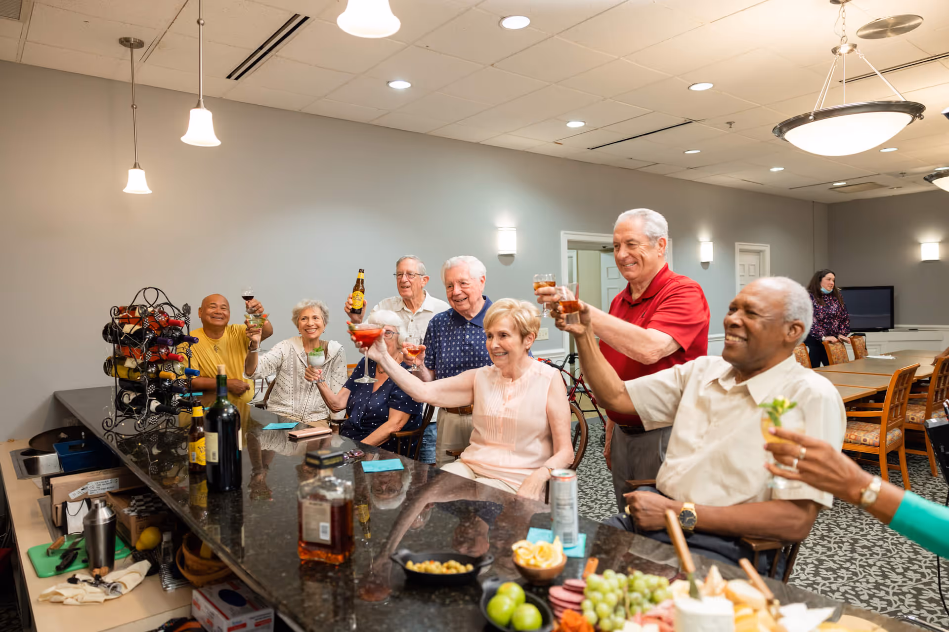 A group of elderly people sitting and standing around a bar counter in a senior living facility, raising their glasses in a toast. The bar counter has various bottles of alcohol, snacks, and a wine rack. The room has a patterned carpet, pendant lights, and a dining area with tables and chairs in the background.