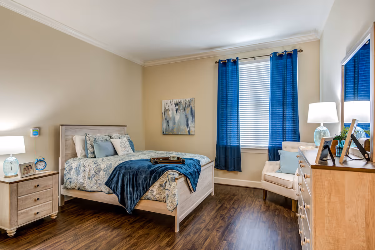 A cozy bedroom with a wooden bed frame, a patterned bedspread in blue and white, and multiple pillows. There is a wooden nightstand with a lamp, a photo frame, and a blue alarm clock. A window with blue curtains and white blinds is on the wall, next to a beige armchair with a blue cushion. A wooden dresser with a lamp, framed photos, and a small plant is also visible. The room has wooden flooring and beige walls.