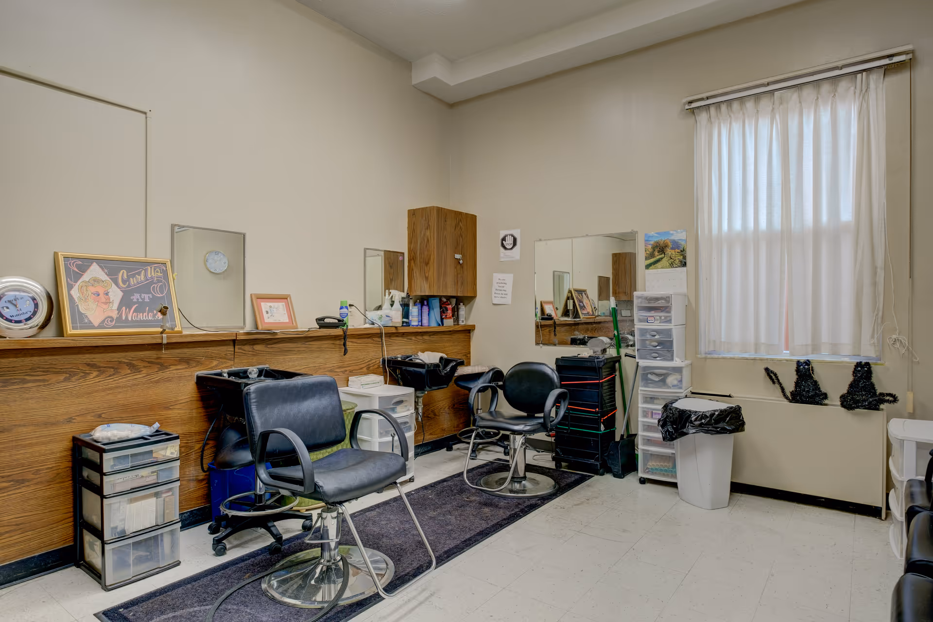 Interior of a small hair salon or barber area with two black salon chairs in front of two hair washing sinks. There are mirrors on the wall above a wooden counter with various hair products. A window with white curtains is on the right side, and several plastic storage drawers and a trash bin are also visible.