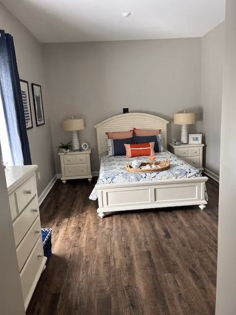 Bright bedroom featuring a white wooden bed with patterned bedding, matching nightstands and dresser, two table lamps, and wood-look flooring.