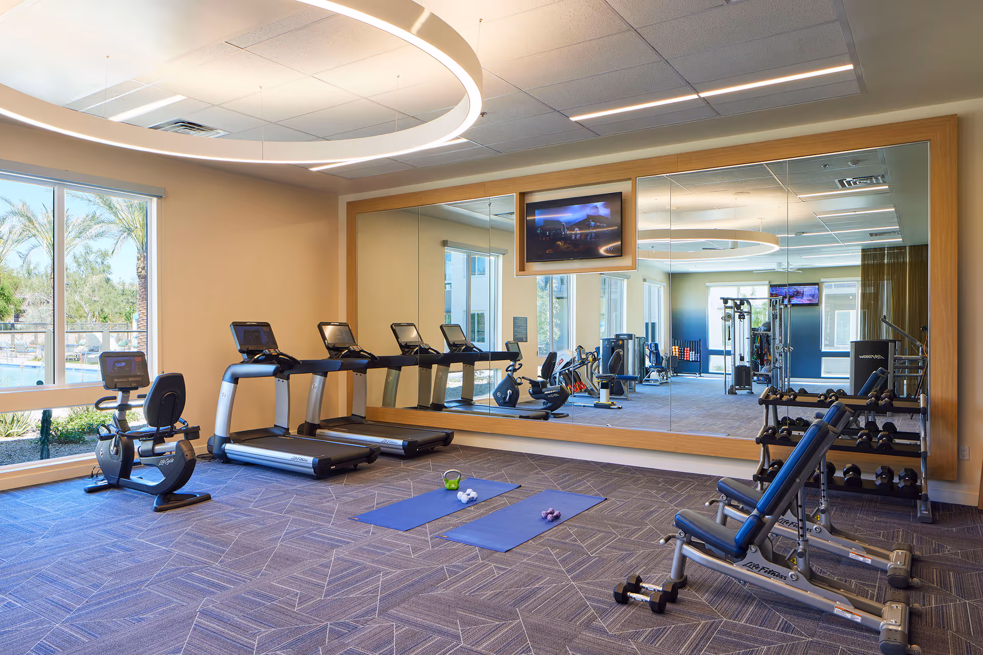 A bright fitness room with exercise equipment including treadmills, stationary bikes, weight benches, dumbbells, and yoga mats. Large mirrors cover one wall, and a flat-screen TV is mounted above the mirrors. Windows show an outdoor pool area with palm trees.