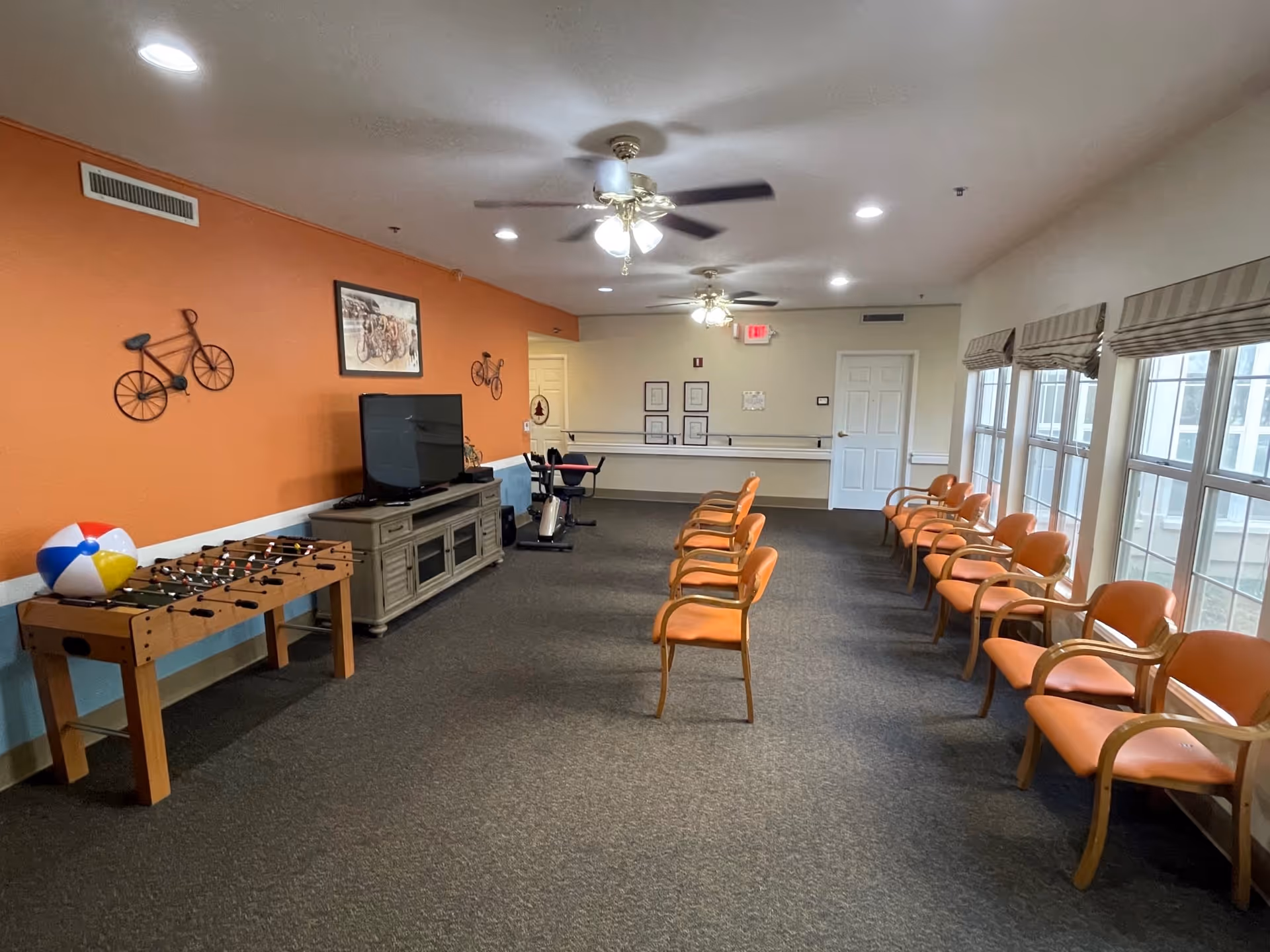 A spacious room with orange walls and multiple orange chairs arranged in two rows facing a TV on a wooden stand. The room has ceiling fans with lights, large windows with blinds on the right side, a foosball table with a beach ball on it, and exercise equipment near the back wall. Bicycle-themed wall decorations are visible on the orange wall.