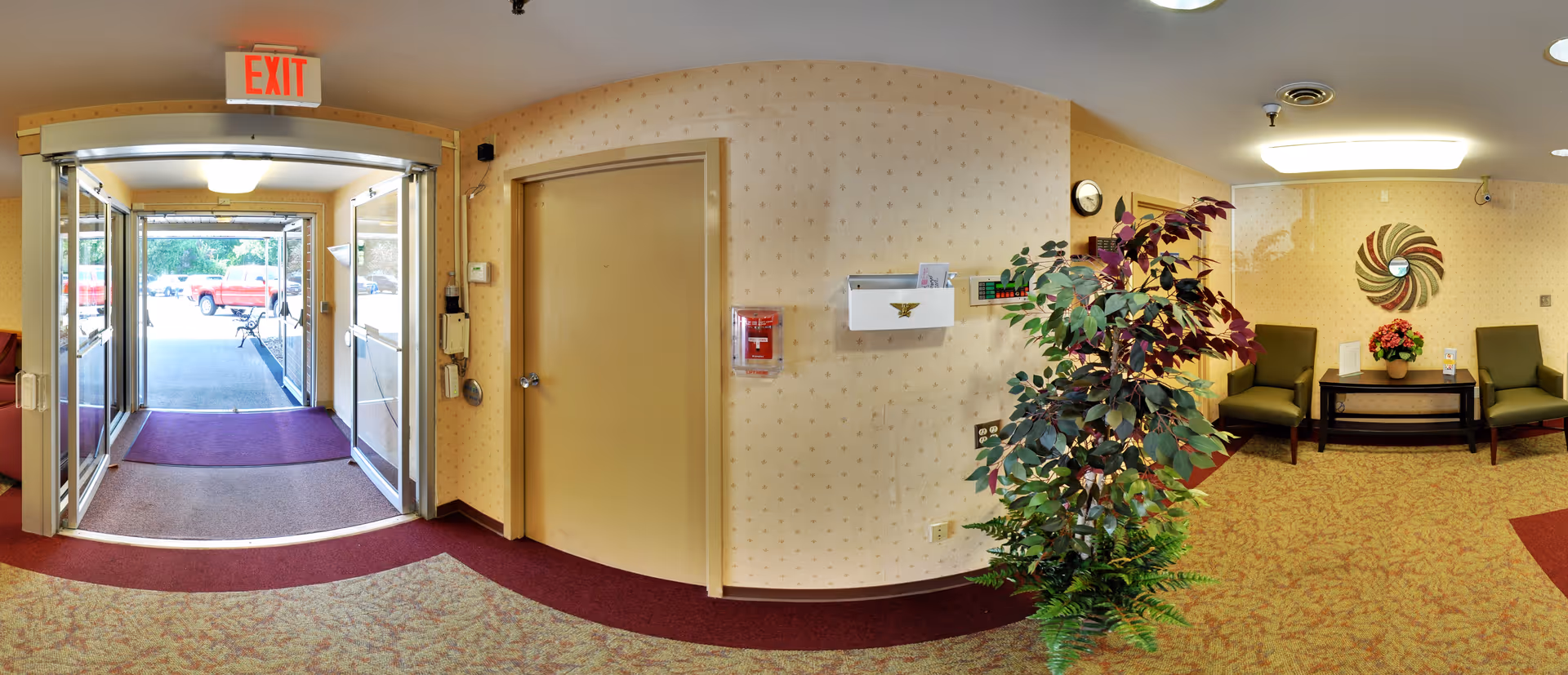 Interior view of a senior living facility entrance area with double glass doors leading outside. The space features patterned wallpaper, a maroon and beige carpet, a green potted plant, two green chairs, a small table with a flower arrangement, a wall clock, and a decorative wall piece. An exit sign is visible above the doors.