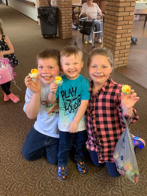 Three smiling children kneeling on a carpeted floor inside a building, each holding a small yellow toy chick. In the background, an elderly person is seated in a wheelchair near brick pillars.