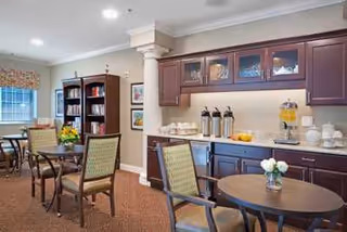 Interior view of a senior living facility common area with round tables and chairs, a dark wood bookshelf filled with books, and a countertop with coffee dispensers, cups, and a beverage dispenser. The room has beige walls, carpeted floor, and a window with a floral valance.