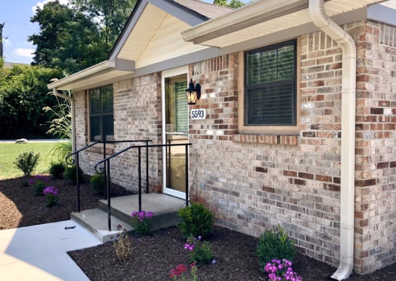 Exterior view of a single-story brick building with a small concrete porch and black metal handrails. The building has two windows with blinds and a door with the number 5593 above it. There are small bushes and purple flowers planted in mulched beds along the front of the building, and a concrete walkway leading to the porch. Trees and greenery are visible in the background under a partly cloudy sky.