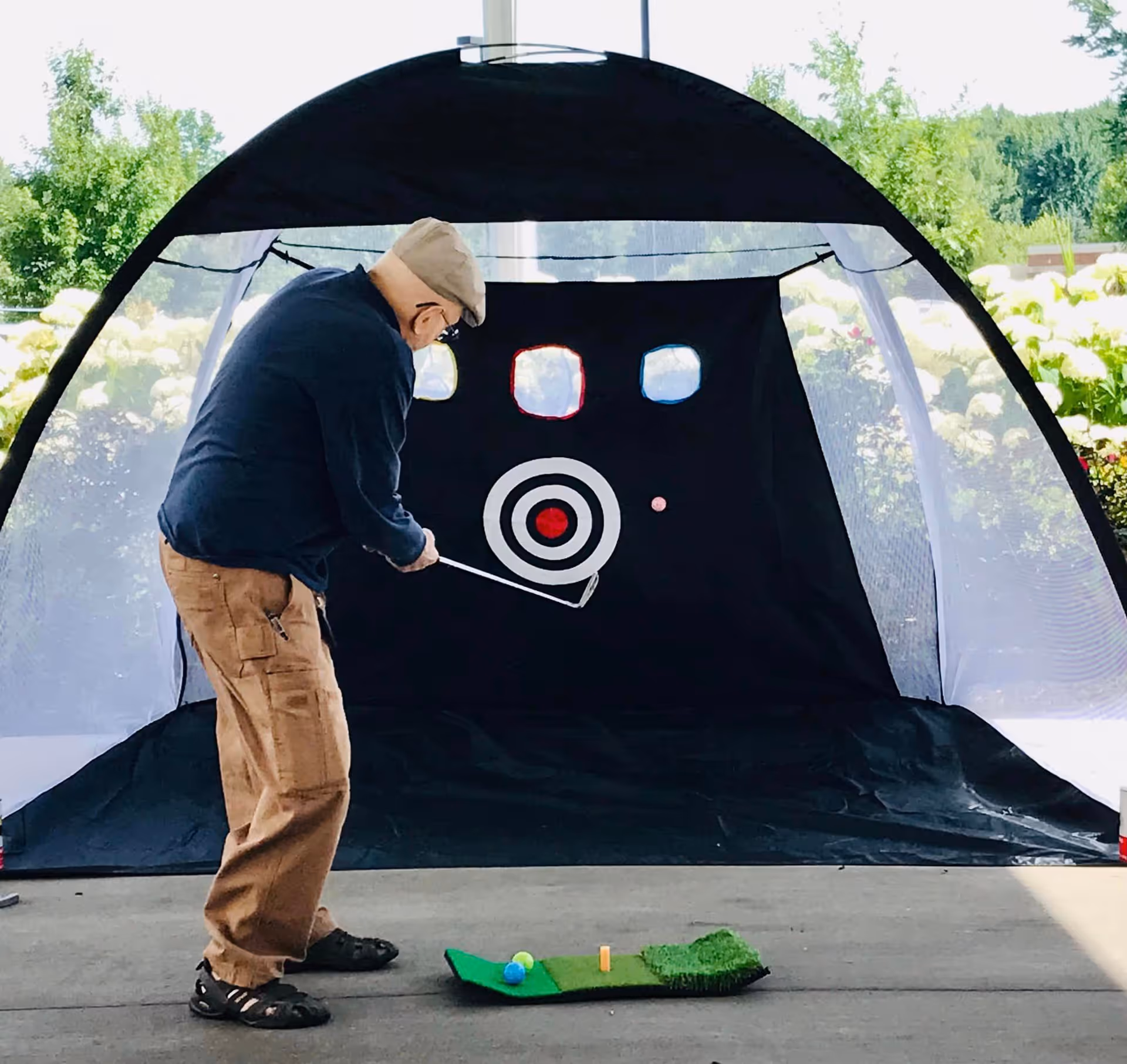 An elderly man wearing a cap, glasses, navy jacket, and tan pants is playing golf indoors using a practice net with a target. There are golf balls and a small green mat on the ground in front of him. The background shows greenery outside through the net.