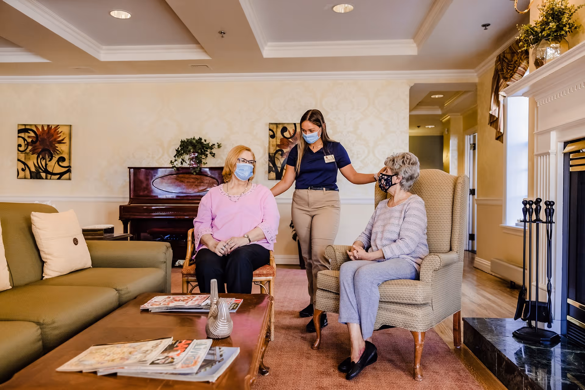 A living room in an assisted living facility with two elderly women sitting and a caregiver standing between them. All three are wearing face masks. The room features a green sofa, a wooden coffee table with magazines, a piano, and a fireplace.