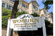 Exterior view of Woburn Rehabilitation & Nursing Center building with a white sign in front displaying the facility name and address, surrounded by trees and greenery under a clear blue sky.