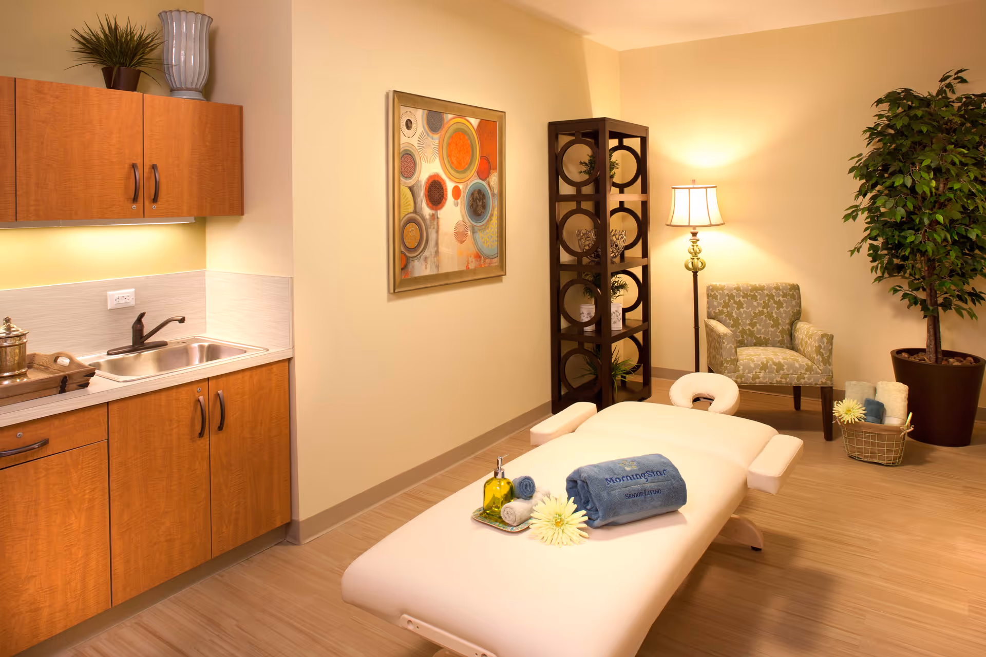 A warm, well-lit massage/treatment room with a massage table, sink and cabinets, an armchair, decorative shelving, artwork and plants.