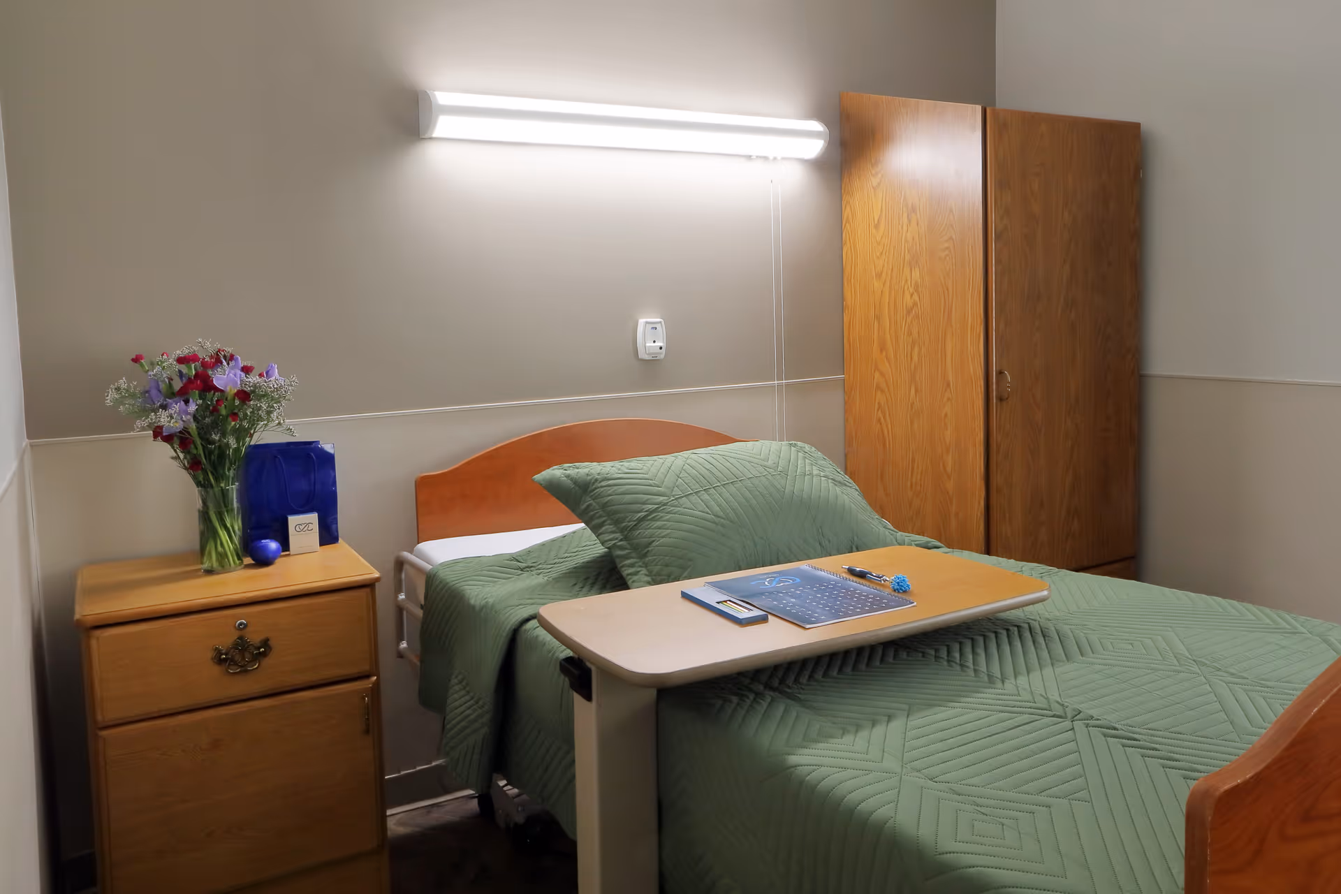 Small senior living bedroom with a single bed covered in a green quilt, an overbed table, bedside drawer with flowers, and a wooden wardrobe.